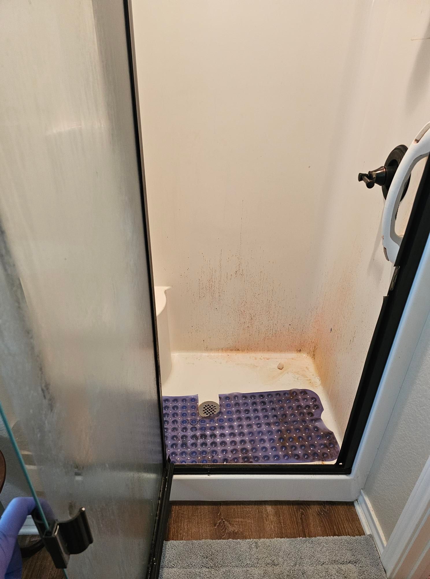 Dirty shower stall with orange stains, a purple mat, and a partially open glass door.
