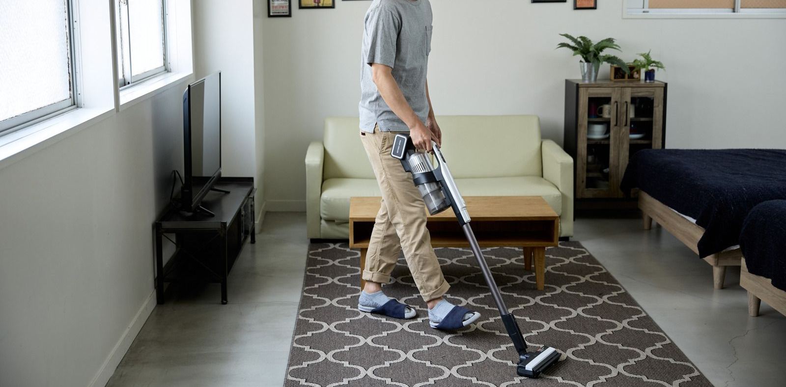 Person vacuums a patterned rug in a well-lit living room with a sofa, coffee table, and a TV.