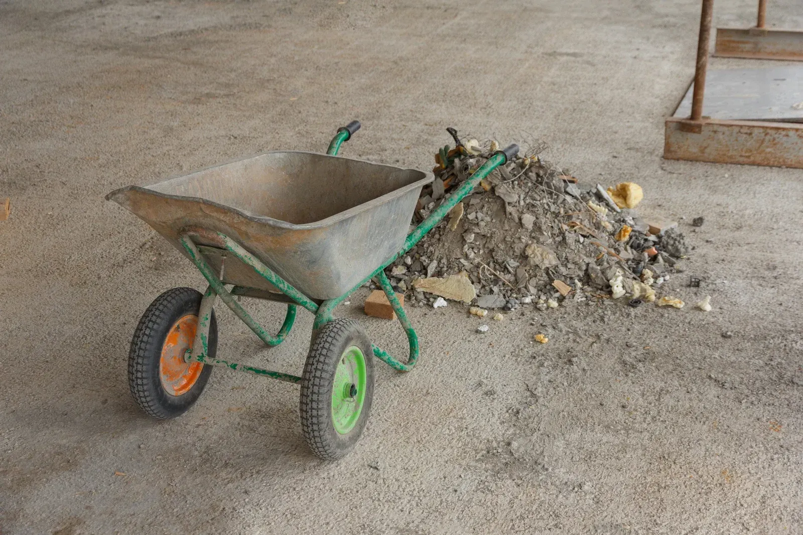 Wheelbarrow with green frame, orange and green wheels, and pile of debris on a concrete surface.