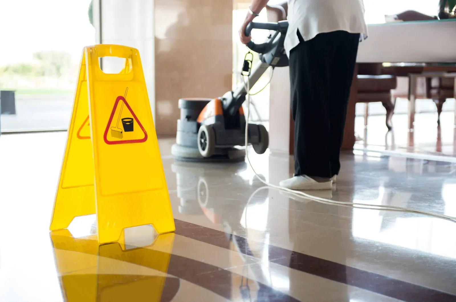 Yellow wet floor sign on shiny floor; person using floor cleaner.