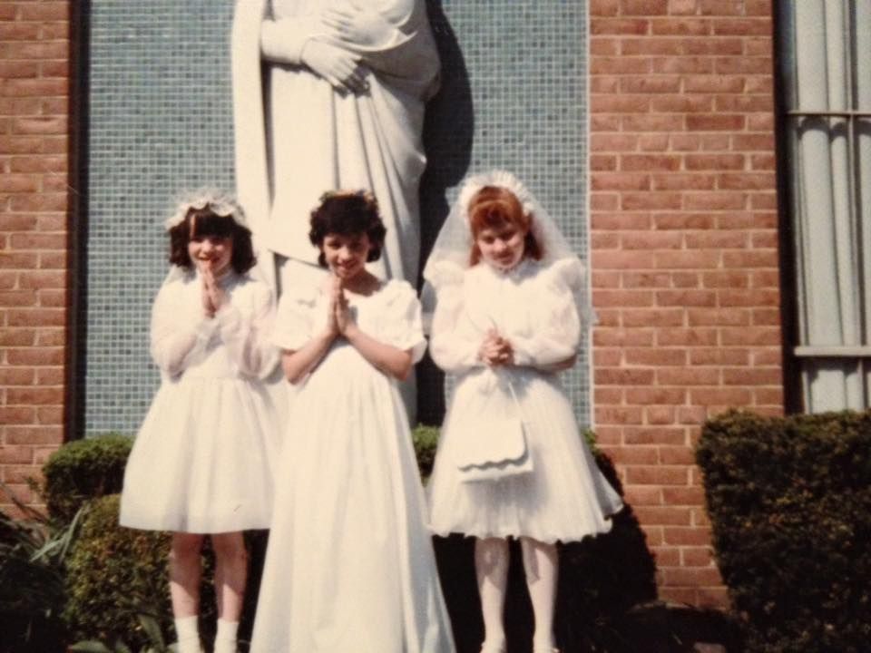 Three girls in white dresses stand in front of a statue