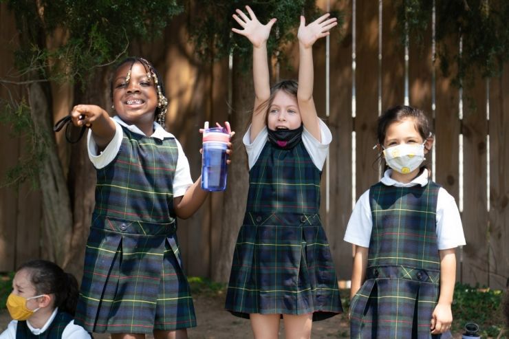 Three young girls wearing face masks and school uniforms are standing in front of a wooden fence.
