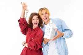 Smiling couple holding a house model, woman in red shirt and man in blue shirt, on a white background