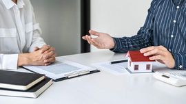 Two people discussing a house model and paperwork at a desk