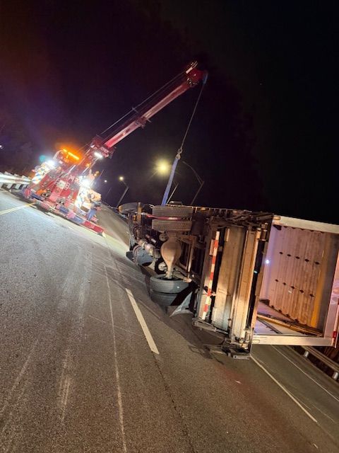 A tow truck uprighting a semi-truck trailer that has overturned on a highway at night.