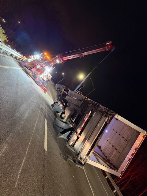 A semi-truck on its side being righted by a tow truck at night on a roadway.