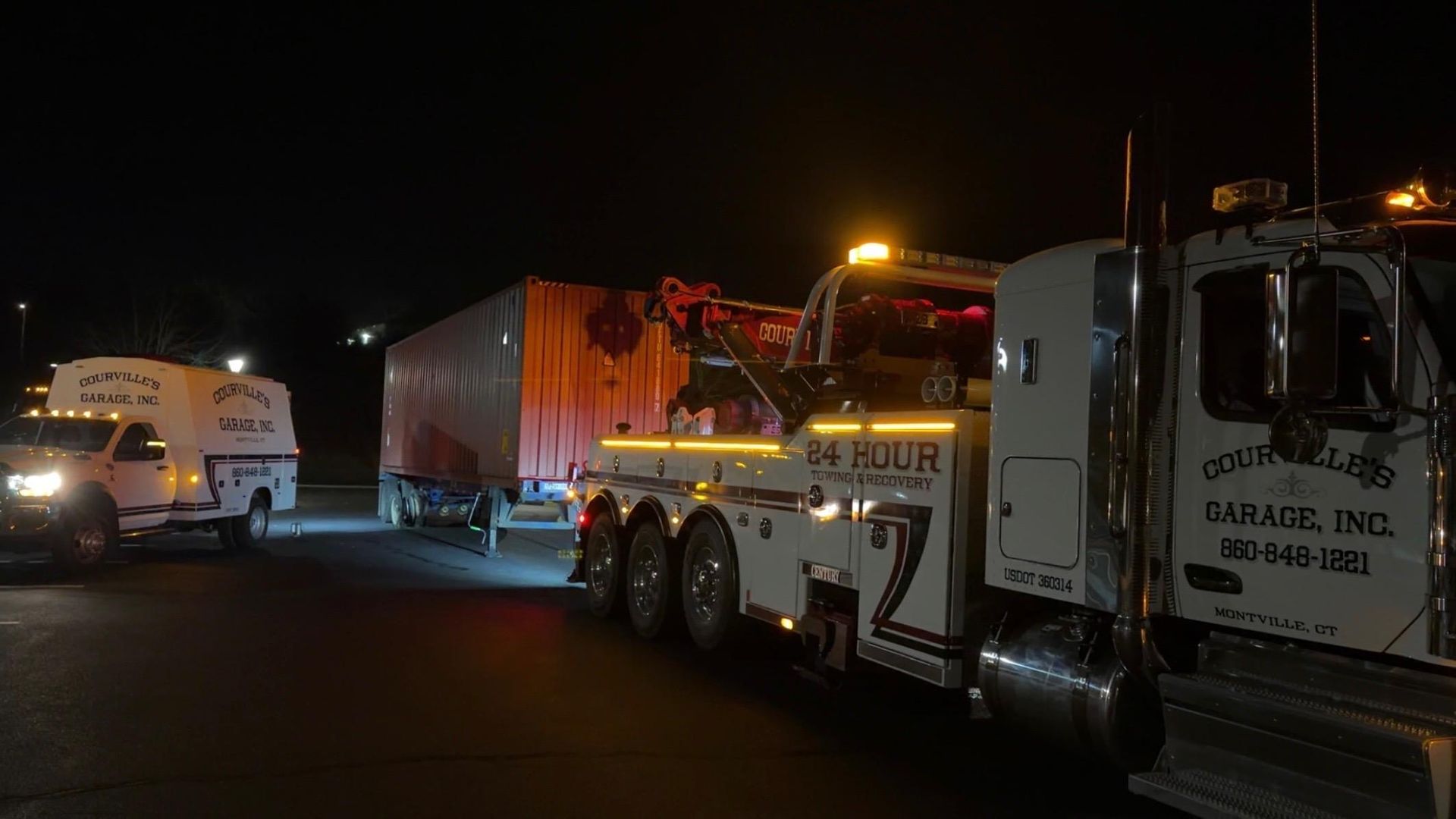 Tow truck towing a large trailer at night with a service truck nearby. Illuminated with flashing lights.