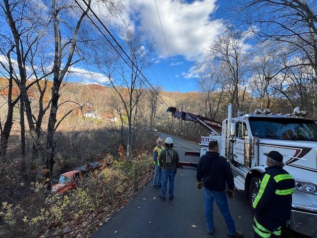 Tow truck retrieving a vehicle from a ditch beside a road; several people observe. Autumn trees, blue sky.