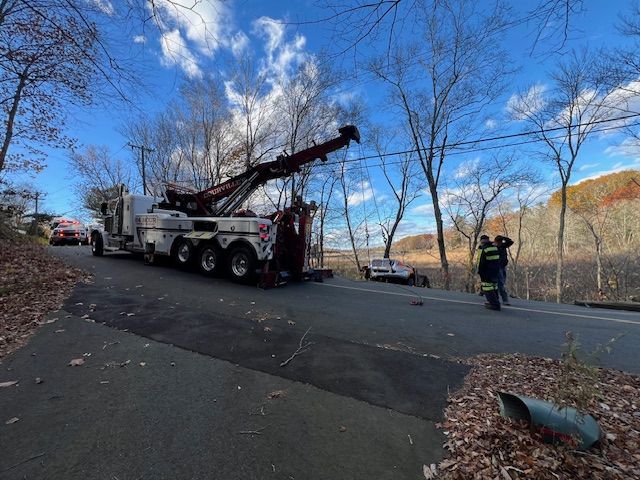 Tow truck removing a car from roadside ditch on a sunny day. Trees in the background, emergency personnel present.