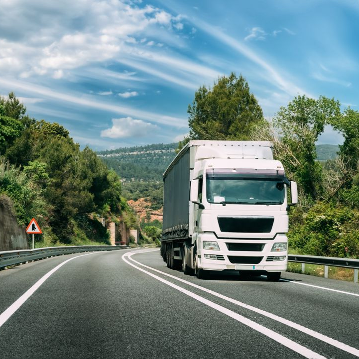 White semi-truck driving on a curving highway under a blue sky with trees and hills