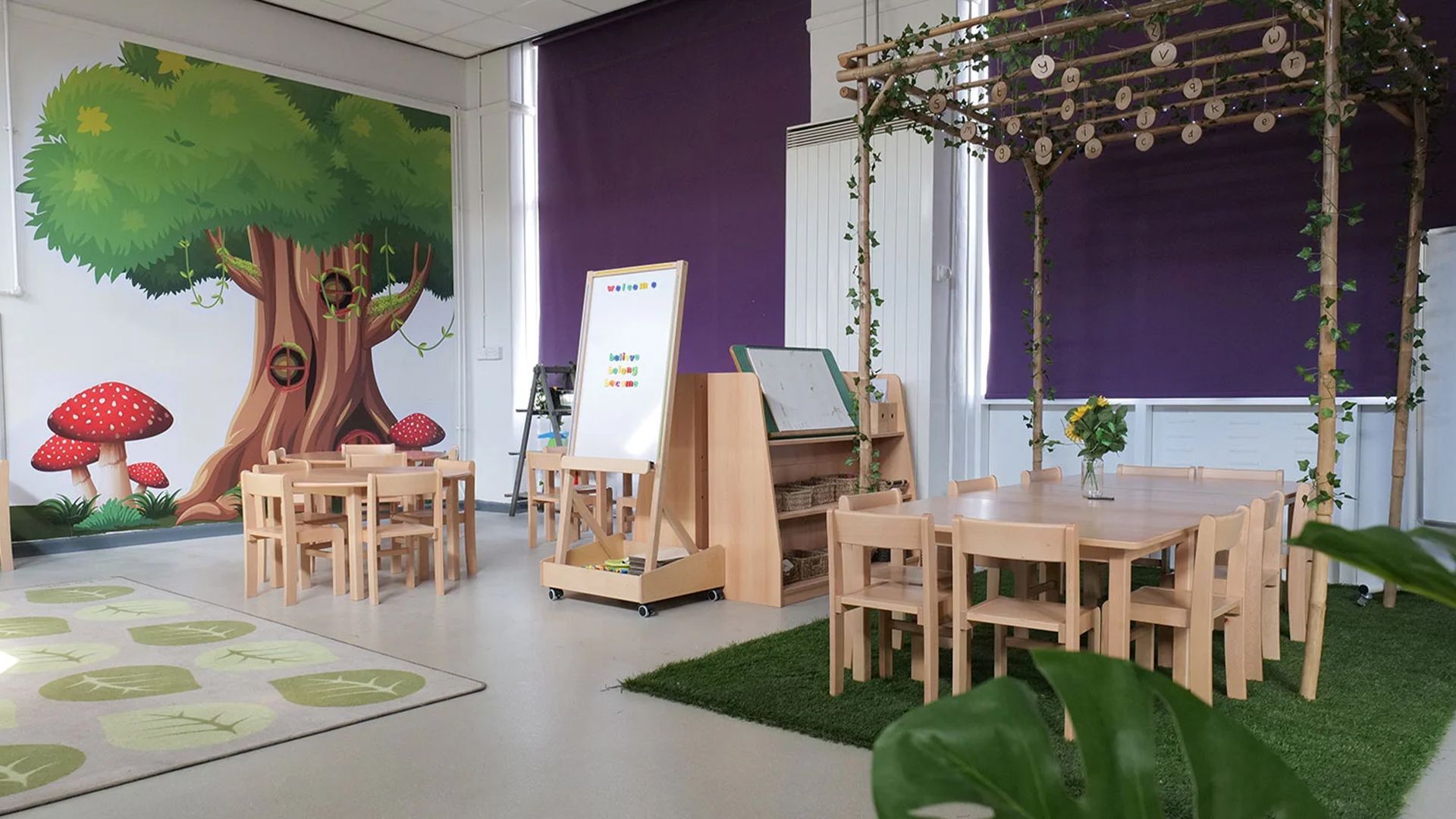 A classroom with tables and chairs and a tree on the wall.