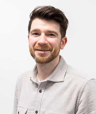 A man with a beard is wearing a grey shirt and smiling for the camera.