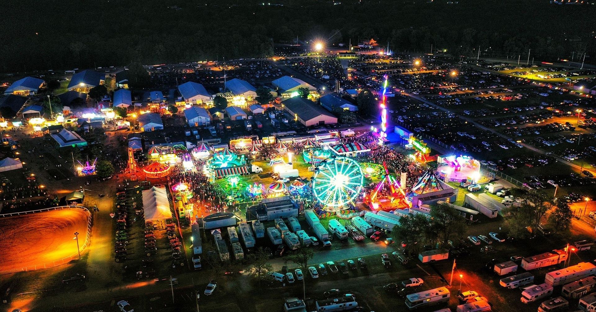 Aerial view of a brightly lit county fair at night, featuring a large Ferris wheel, carnival rides, and rows of campers.
