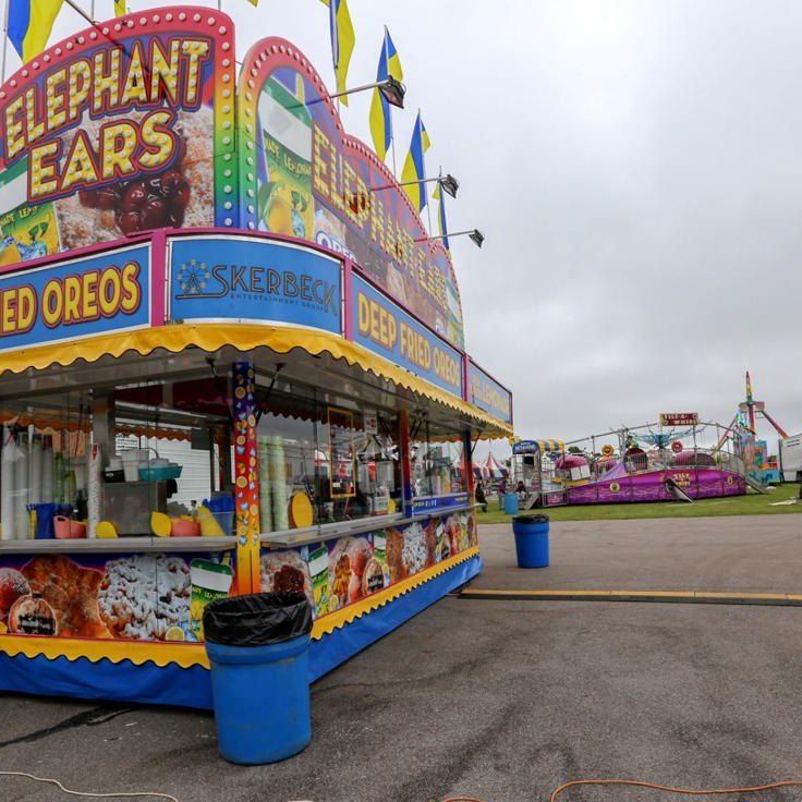 A colorful carnival food stand selling elephant ears, deep-fried Oreos, and other treats, with fair rides in the distance.