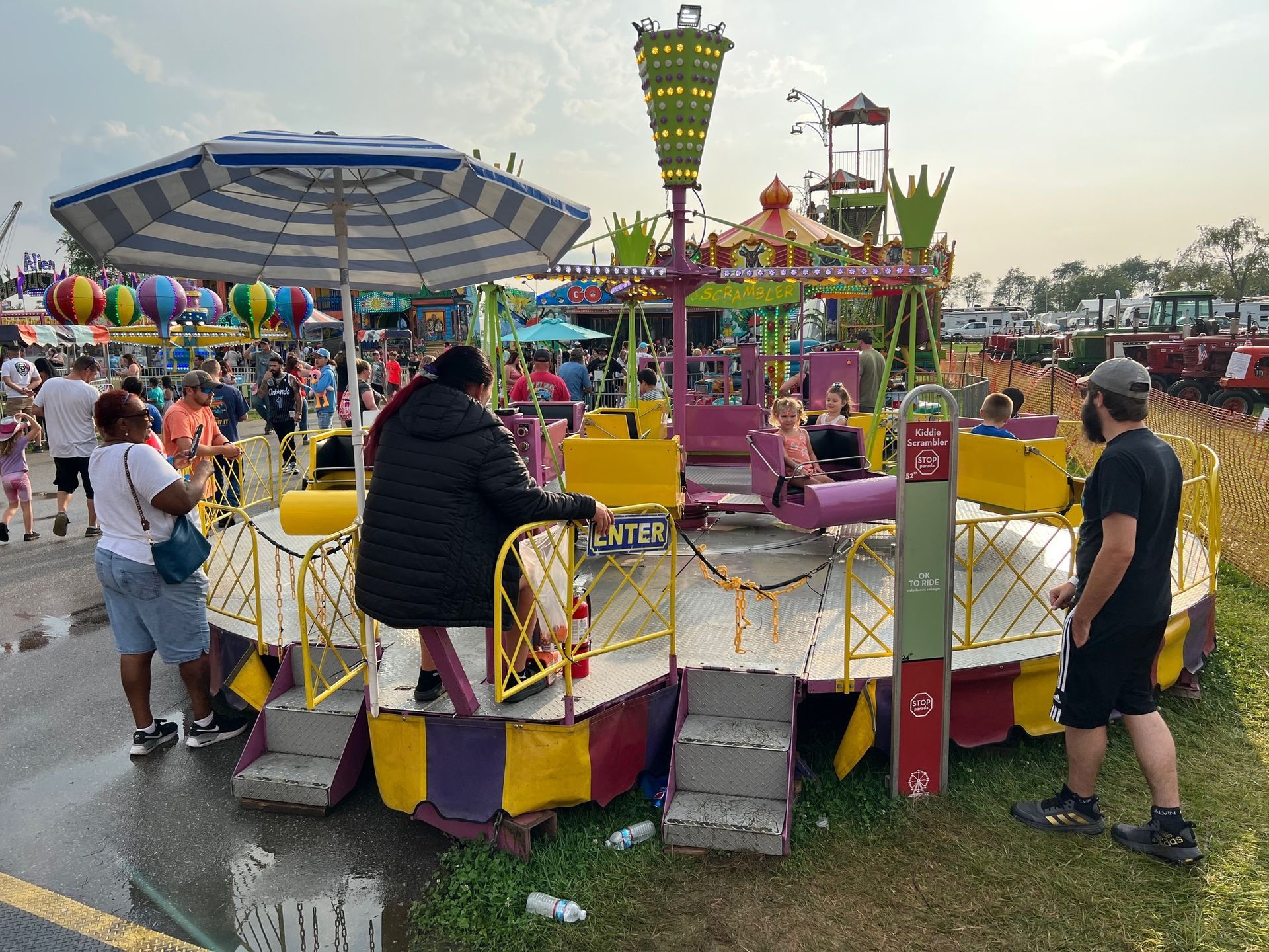 People stand near a fairground carousel ride under a large striped umbrella on a sunny day.