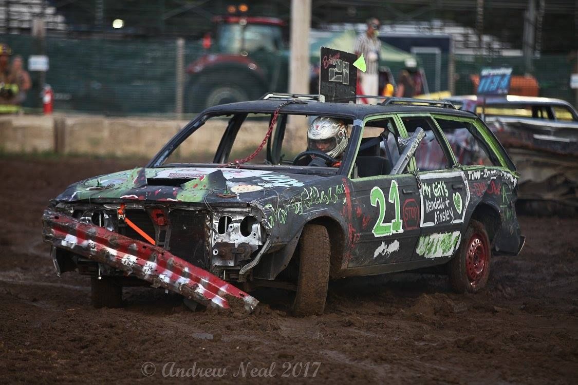 A black demolition derby station wagon with the number 21 painted in green, driving on a muddy dirt track.