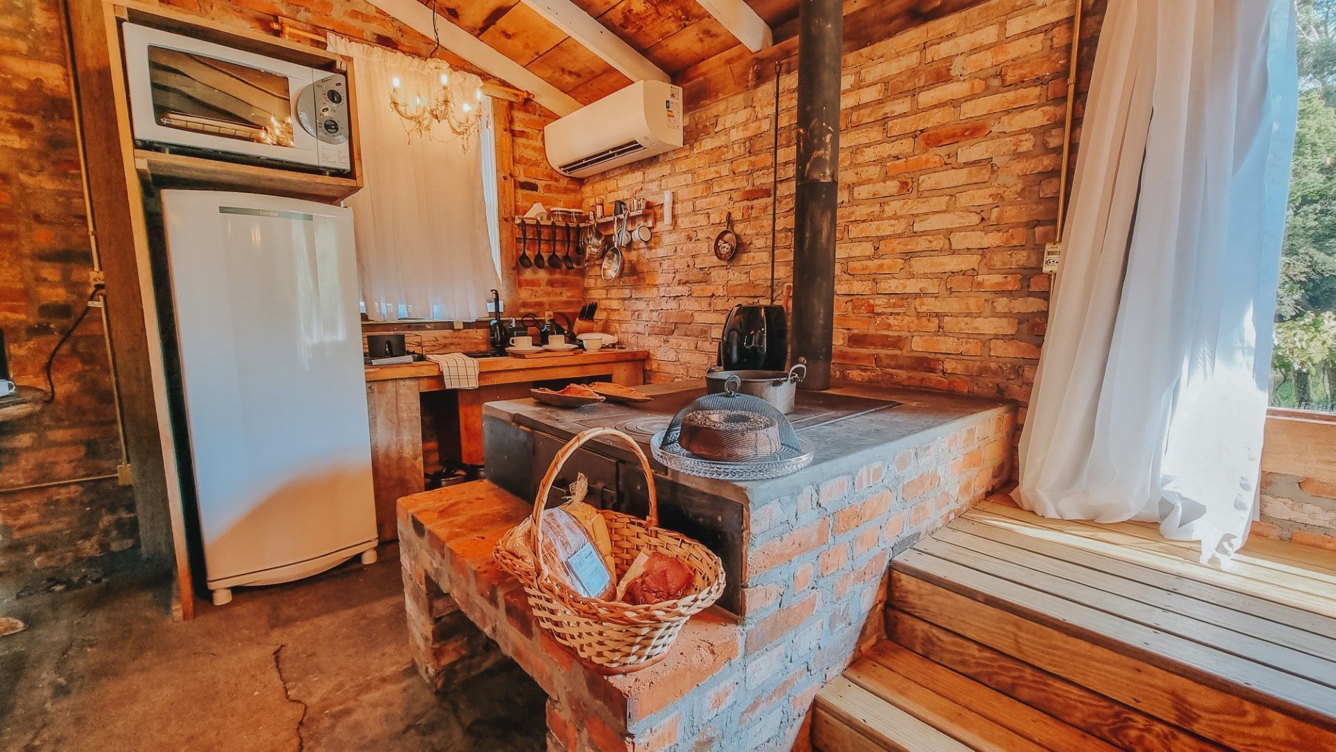 Cozinha rústica com paredes de tijolos e um fogão de pedra. Uma cesta de pães repousa na borda do fogão.