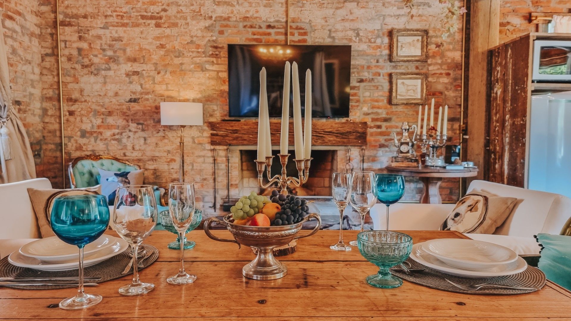 Mesa de jantar formal posta para convidados em uma sala com tijolos aparentes e lareira. Copos azul-petróleo e velas.