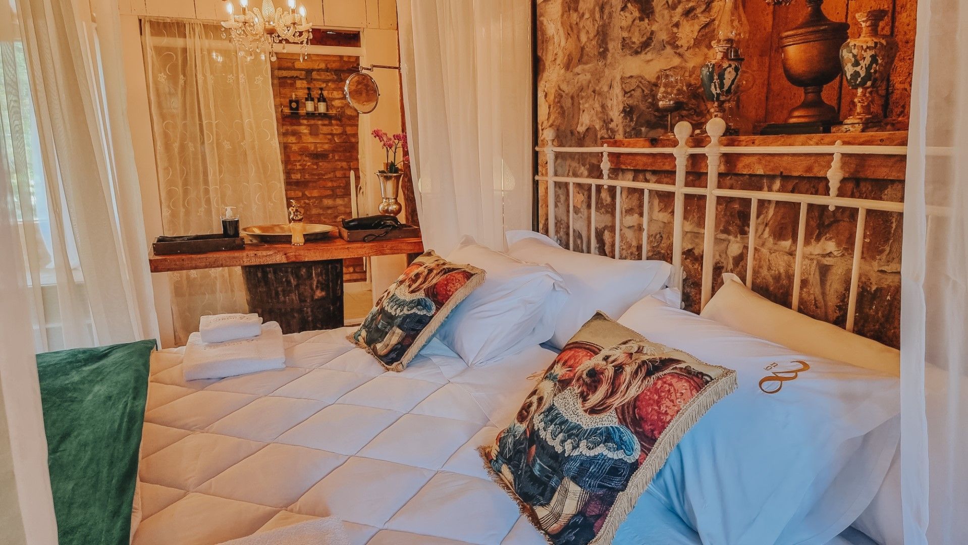 Quarto aconchegante com cama branca, almofadas ornamentadas e cortinas transparentes. Escrivaninha de madeira e parede de pedra adicionam um charme rústico.