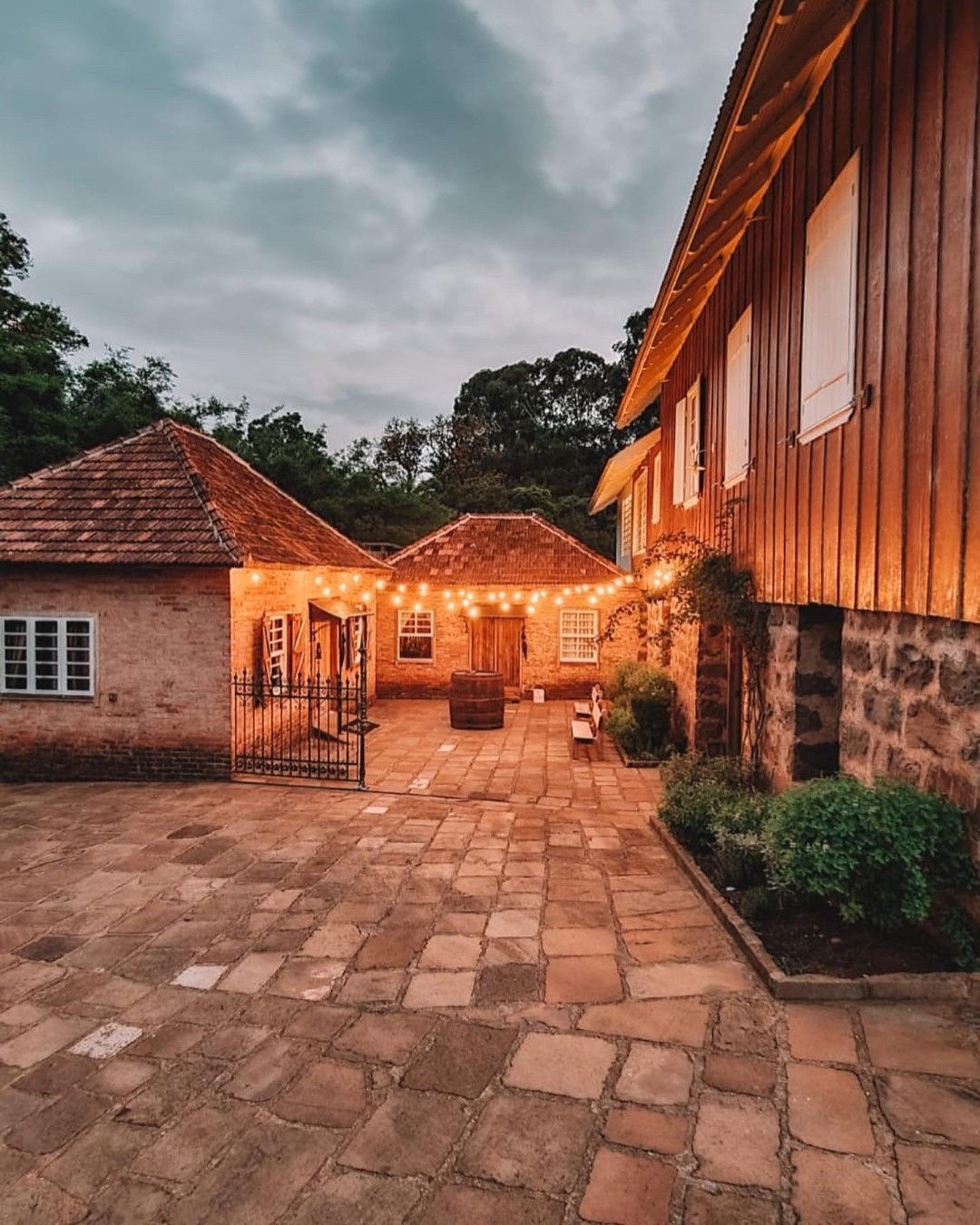 Pátio com construções de tijolos e madeira, iluminado por luzes de corda, ao entardecer.