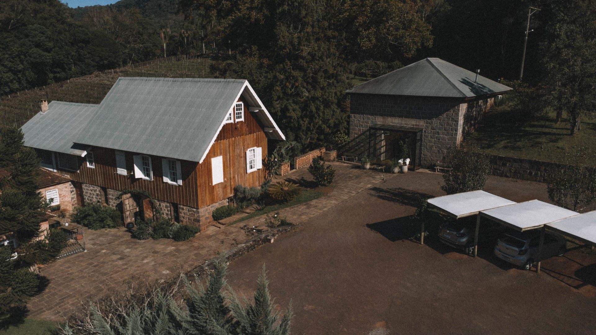 Casa de madeira e edifício de pedra com estacionamento, situados em uma área arborizada.