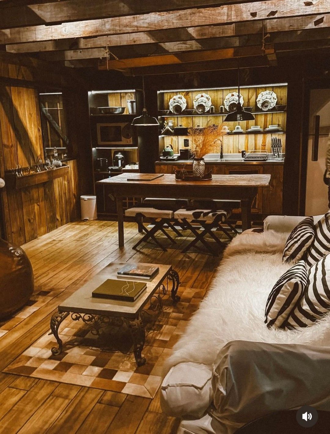 Interior aconchegante de cabana com paredes e pisos de madeira. Dispõe de cozinha, mesa de jantar e área de estar com tapete e sofá.