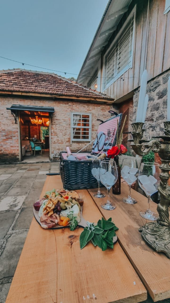Mesa de madeira posta para um piquenique ao ar livre, com frios, taças de vinho e uma cesta, perto de um prédio de tijolos.