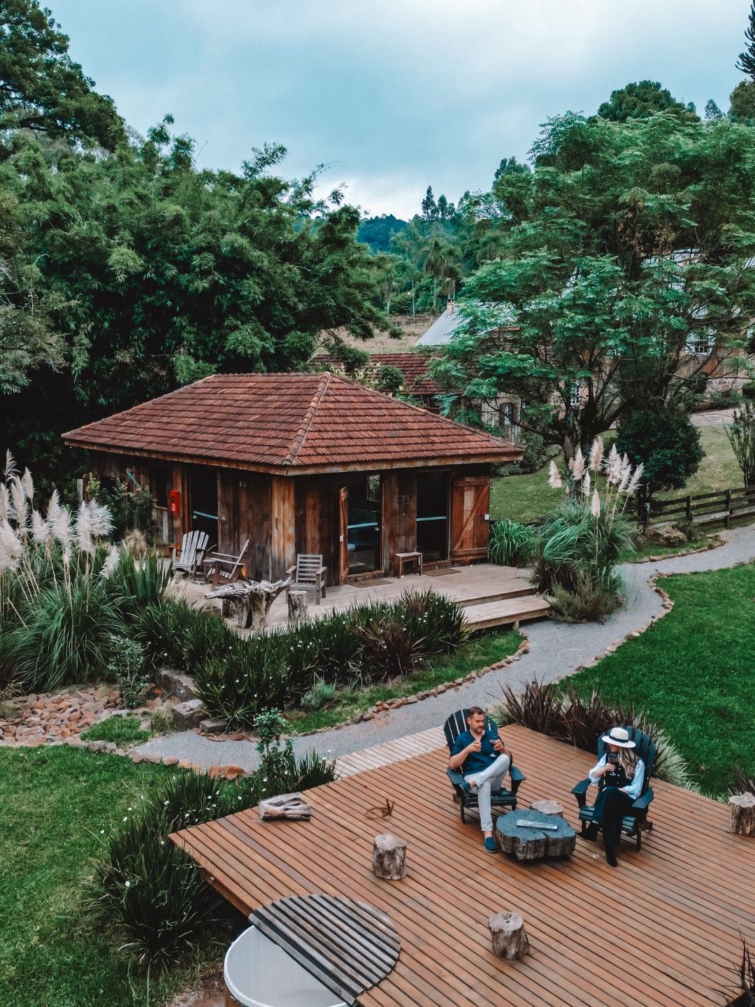 Cabana de madeira com deck, cercada por árvores; duas pessoas sentadas no deck, montanhas visíveis ao longe.