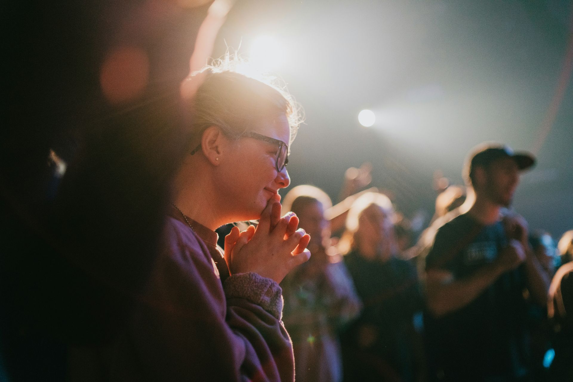 Woman with glasses watches, hands clasped, at a concert with bright stage lights.