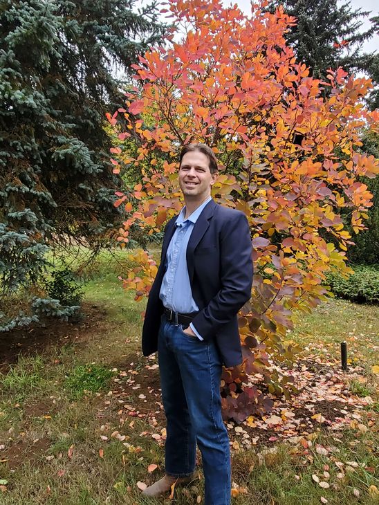 Darrel is standing outdoors in front of  a small tree.