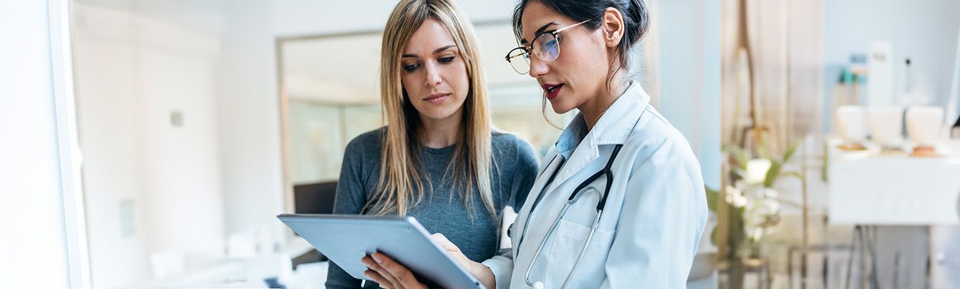 A clinician in a white coat explains medical information on a tablet to a patient in a bright, modern office.