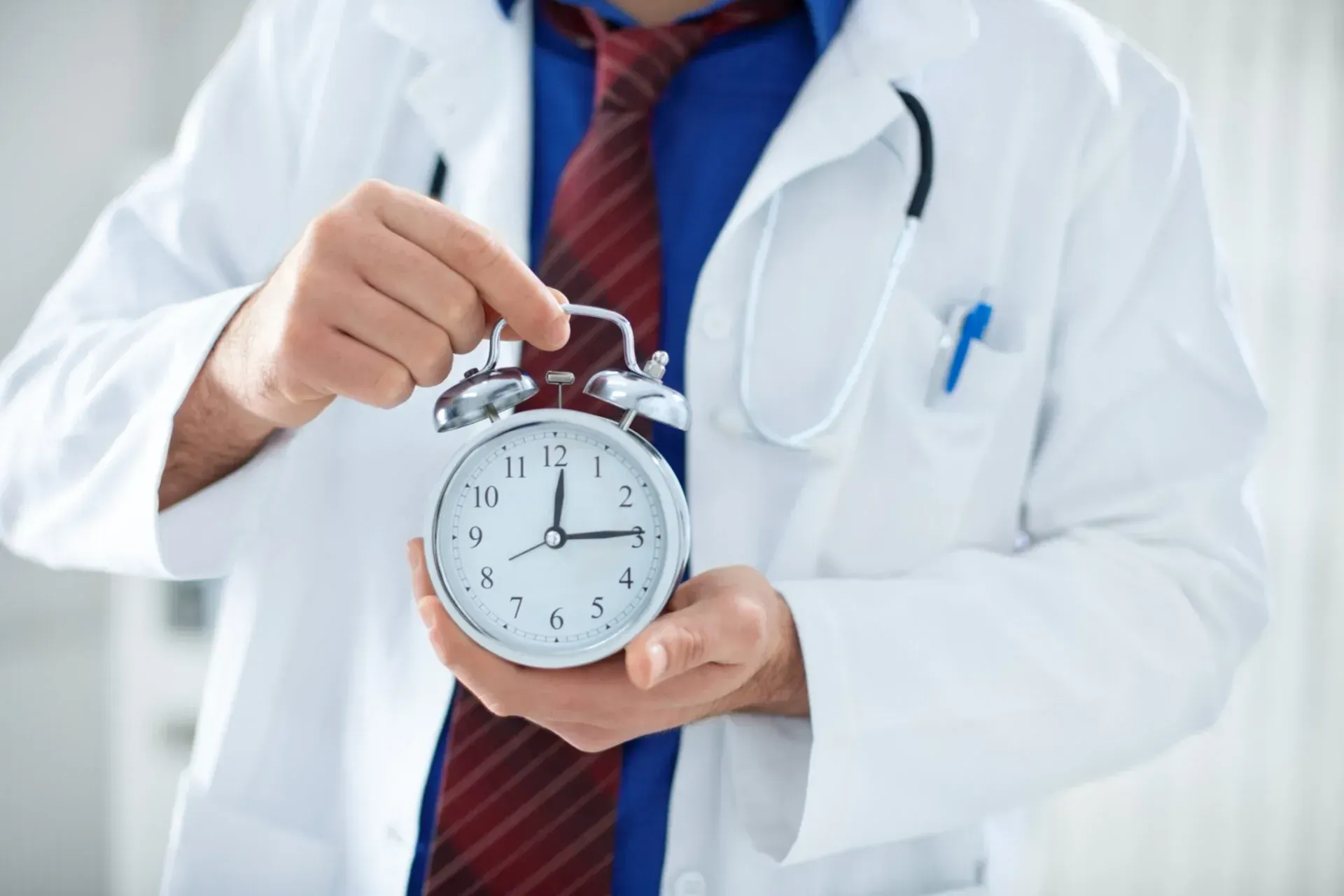 A person in a white doctor’s coat and blue shirt holding an analog alarm clock.