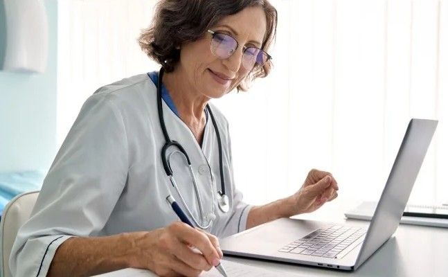 Doctor, wearing glasses and stethoscope, taking notes while using a laptop in an office.
