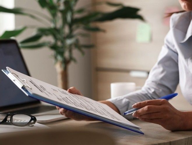 Person holding a clipboard, reviewing documents, pen in hand, near a laptop and plant.