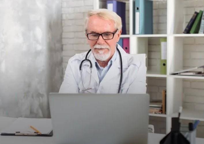 Doctor at desk, looking at laptop. White coat, stethoscope, office setting with bookshelf.