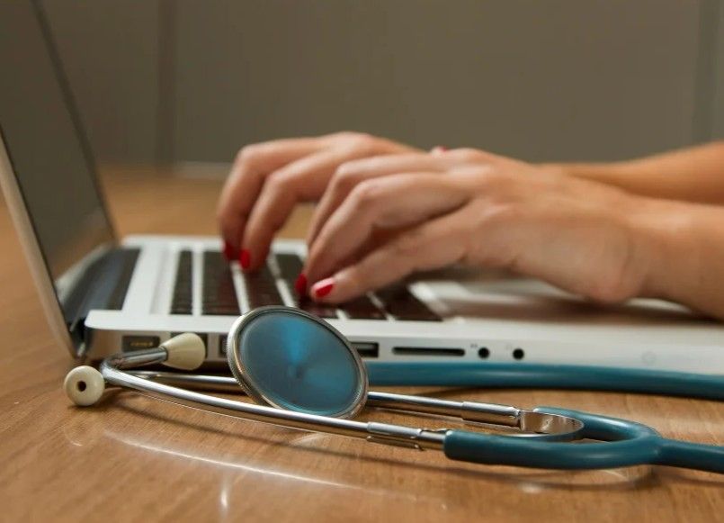 Hands typing on a laptop with a stethoscope lying in the foreground.