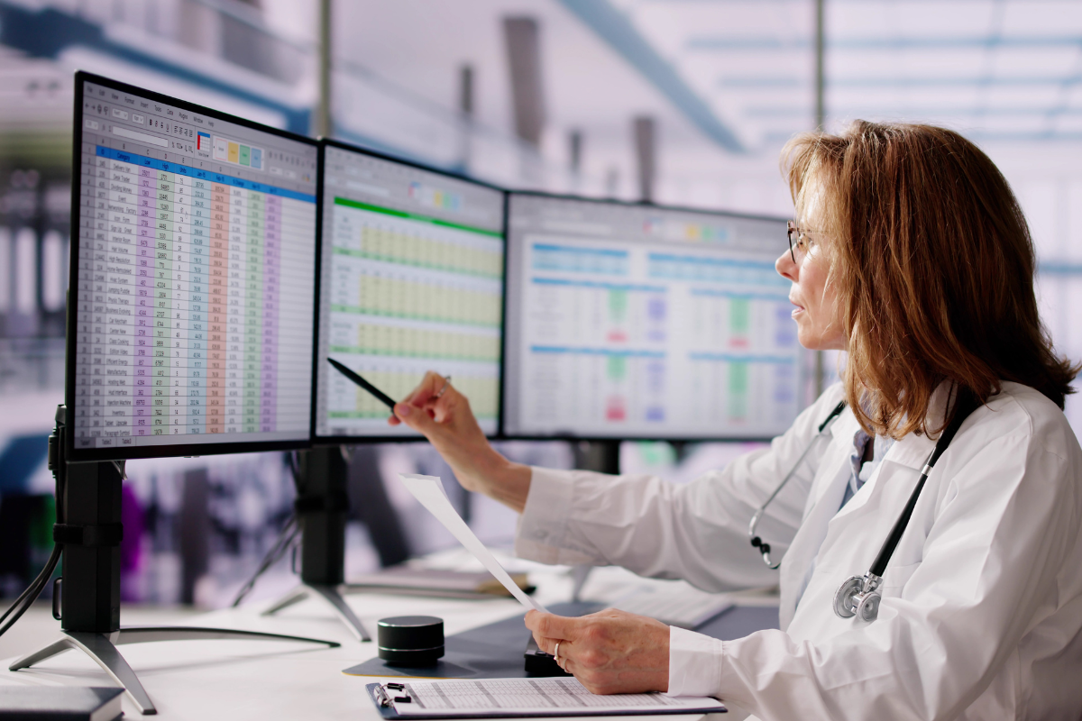 Doctor in white coat pointing at data on three computer screens.