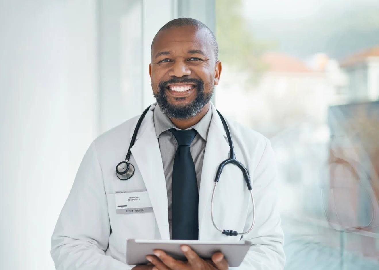Doctor in white coat smiles, holds tablet, stethoscope around neck, standing near window.