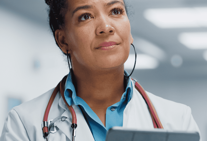 Doctor in white coat, stethoscope, holding tablet, looking upward with thoughtful expression in a hospital hallway.