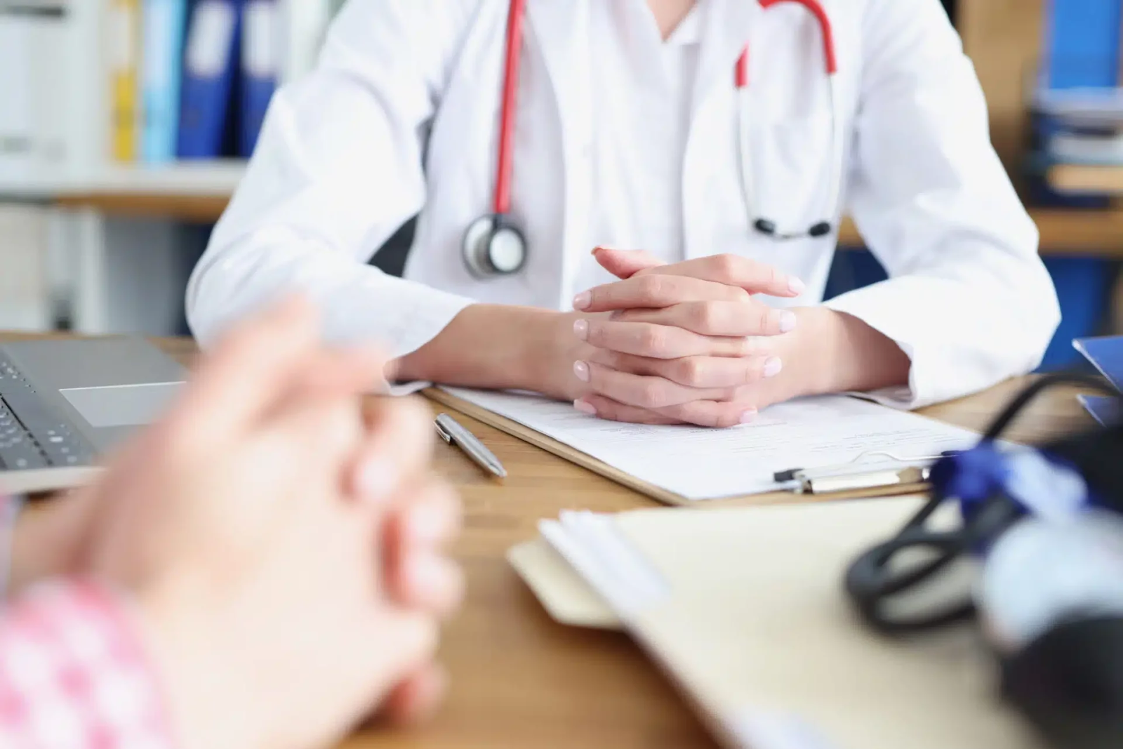 Doctor with stethoscope at desk, hands clasped, facing patient. Clipboard and medical equipment visible.