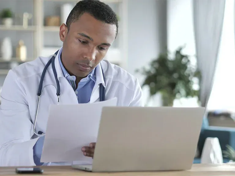 A person in a white lab coat with a stethoscope around their neck reviewing documents while sitting at a laptop.