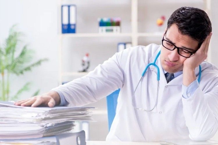 Doctor in a white coat looking exhausted, resting head on hand. Papers on desk, stethoscope around neck.