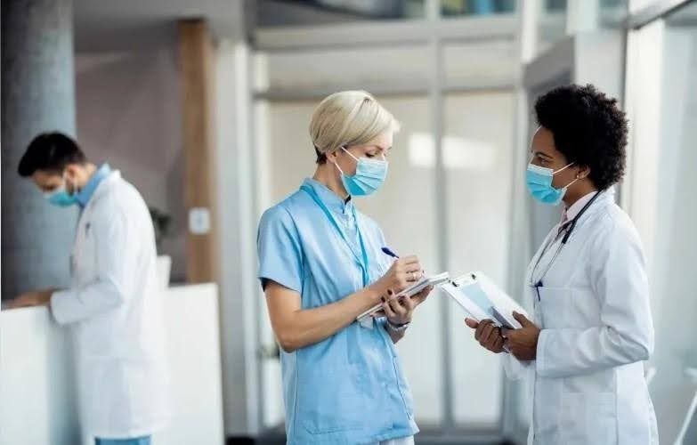 Medical professionals in a hospital hallway wearing masks; one writing notes, another holding a clipboard.
