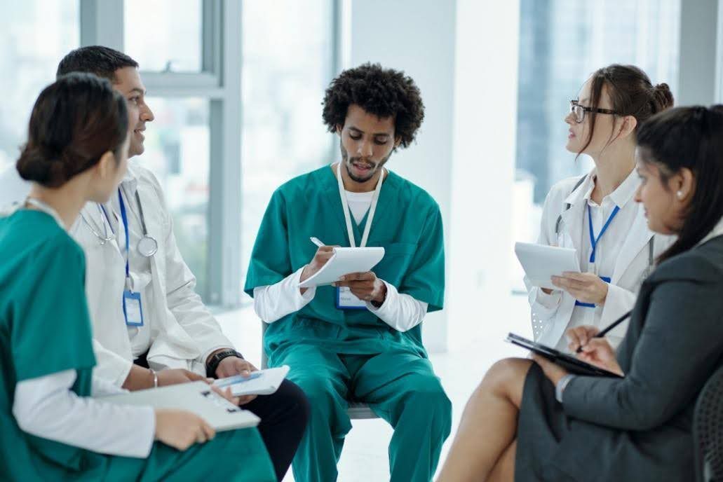 Medical team in scrubs and lab coats meeting, taking notes, light-filled room.