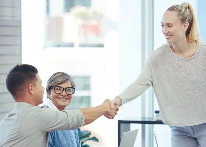 Three people in a bright office space shake hands, smiling as they meet.