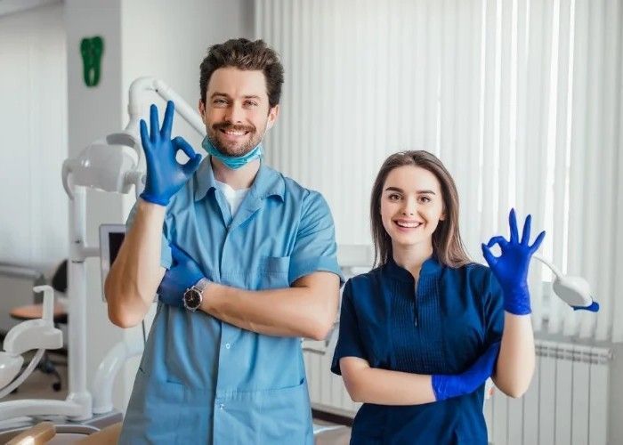 A dentist and assistant in scrubs with blue gloves make an 'okay' gesture in a dental office.