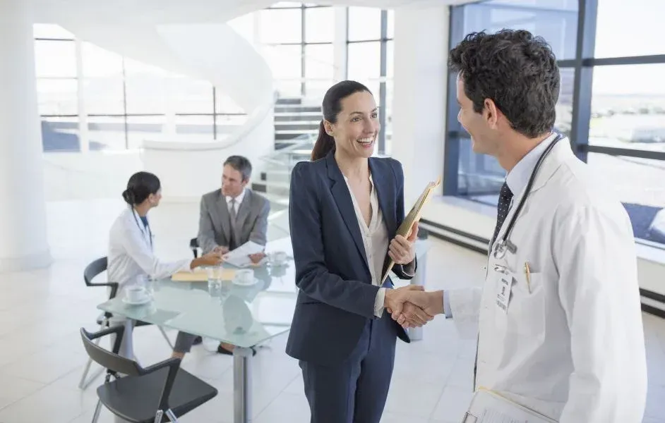 A professional in a suit shakes hands with a medical professional in a lab coat inside a bright, modern office.