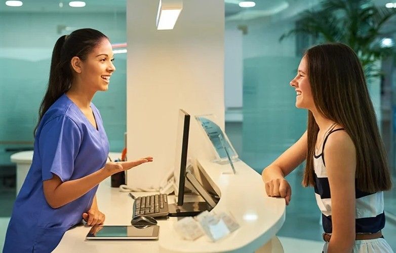 Receptionist in blue scrubs smiles and talks to a patient at a white counter.