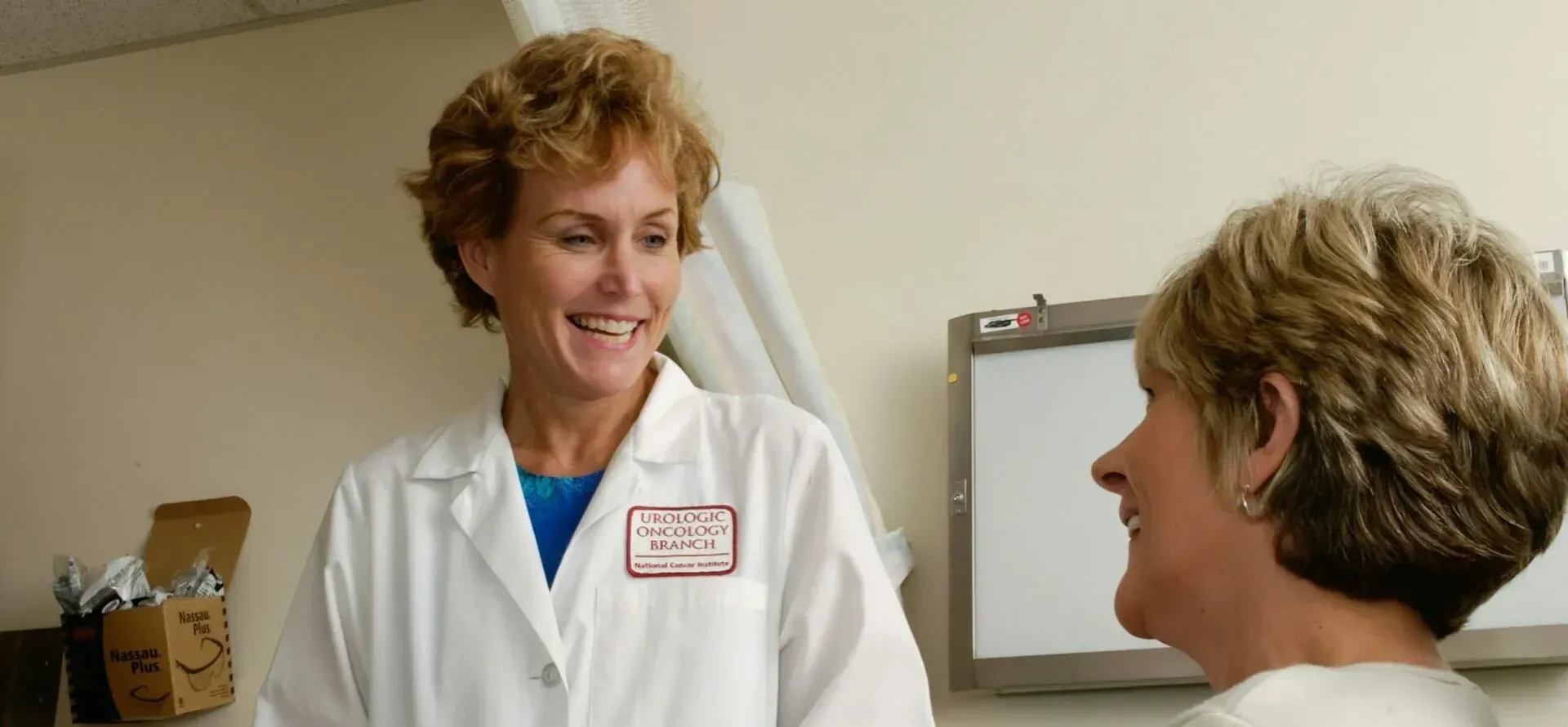 Doctor in white coat smiling, talking to a patient in an examination room.