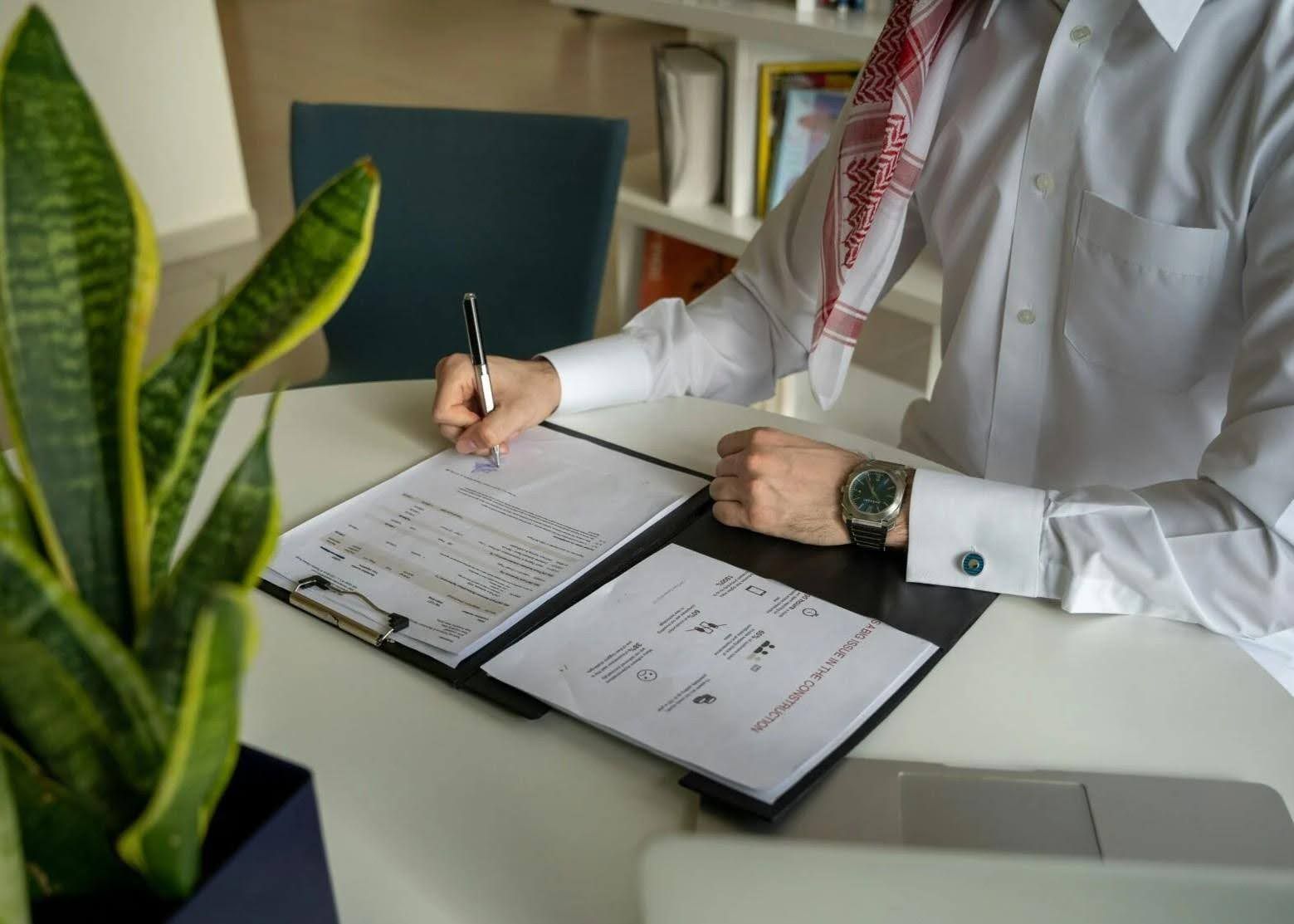 Person in traditional headwear writing on papers at a desk with a plant.